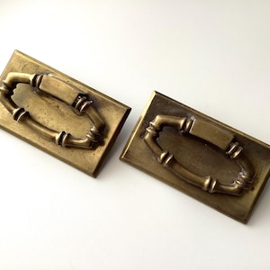 May include: Two antique brass drawer pulls with a rectangular backplate and an oval handle. The handles have a bamboo-like design, adding a touch of vintage charm. The brass has a warm, aged patina.
