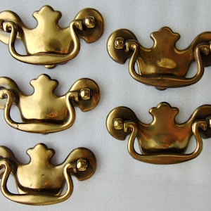 May include: Five antique brass drawer pulls with a decorative crown-shaped top and a curved handle. The pulls are in excellent condition and have a beautiful patina.