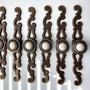 May include: Six antique bronze cabinet pulls with a decorative design. Each pull features a round, off-white center surrounded by a black and bronze beaded border. The pulls have a scrolling, elongated shape.