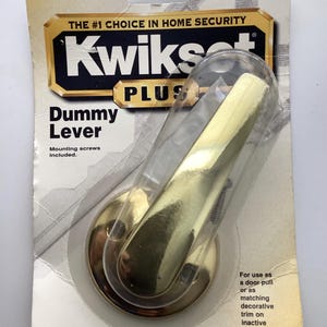May include: A Kwikset Plus dummy lever door handle in a clear plastic package. The handle is gold-colored and has a curved lever design. The packaging includes the text "THE #1 CHOICE IN HOME SECURITY" and "Dummy Lever".