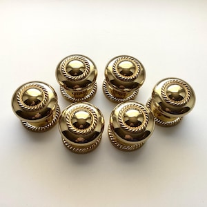 May include: Six gold-colored cabinet knobs with a decorative rope-like design around the edges. The knobs have a rounded shape and a shiny finish, suggesting a classic or vintage style. They are arranged on a white surface.