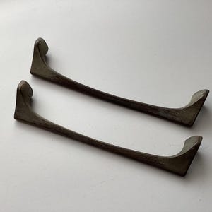 May include: Two dark brown, curved wooden shelf brackets. Each bracket has a long, slender arm with a curved end. The brackets are positioned on a white surface, showcasing their design and form.