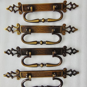 May include: Four antique bronze drawer pulls with ornate detailing. Each pull features a rectangular backplate with a textured surface and decorative end pieces. The handles are curved and have a central decorative element, perfect for vintage furniture restoration.