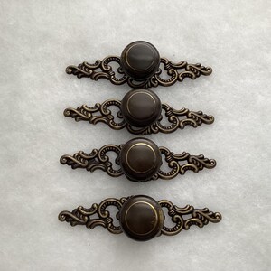 May include: Four drawer pulls featuring a dark brown circular knob centered on an ornate, antique brass-toned metal backplate with intricate scrollwork detailing. Each pull has a thin gold-colored ring accent around the top of the knob.