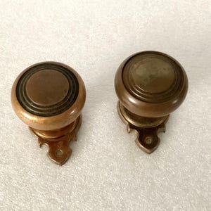 May include: Two antique bronze cabinet knobs. Each knob has a round, tiered design with a decorative base. The knobs are a warm, aged bronze color, suggesting they are vintage or have a classic aesthetic. They are likely used for furniture or cabinetry.