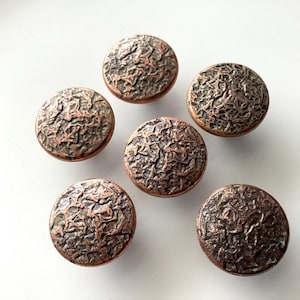 May include: Six round, textured copper-colored cabinet knobs. The knobs have a detailed, organic pattern across their surface, giving them a rustic appearance. They are arranged on a white surface.
