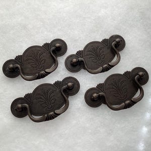 May include: Four dark bronze-toned metal drawer pulls featuring a rounded backplate with an engraved floral pattern and a curved, swinging handle. Each piece has a symmetrical design with circular ends and a central decorative motif.