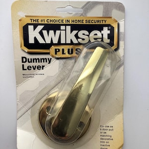 May include: A Kwikset Plus dummy lever door handle in a clear plastic package. The handle is gold-colored and has a curved lever design. The packaging includes the text "THE #1 CHOICE IN HOME SECURITY" and "Dummy Lever".