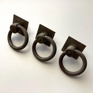 May include: Three antique bronze ring pulls with square backplates. The ring pulls are circular and attached to the square backplates with a cylindrical base. The hardware has a weathered, aged appearance.