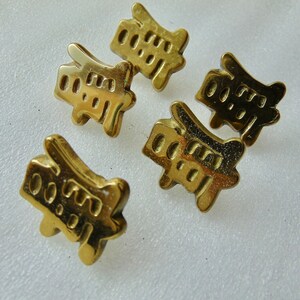 May include: Five small, gold-colored metal pins. Each pin has a unique shape with a series of cut-out shapes and holes. The pins are arranged on a white surface, with the light reflecting off the shiny metal.