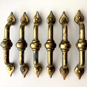 May include: Six antique brass door handles with a curved design. Each handle features a decorative top and bottom, with a central bulbous section. The brass has a vintage patina, showing signs of age and use, adding to their character.