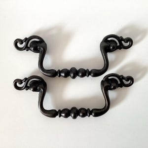 May include: Four antique bronze-colored drawer pulls. Each pull has a curved handle with decorative scrollwork on the ends and a series of rounded beads in the center. The pulls are set against a white background.