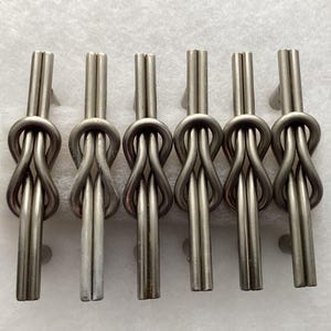 May include: Six silver-toned metal tie clips arranged in a row. Each clip features a knot design at the center, with a vertical bar extending upwards. The clips are displayed on a white surface.