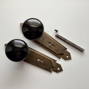 May include: Two antique door knobs with black glass spheres, mounted on brass plates with keyholes. A small screw and a metal bar are also present, suggesting a restoration project or hardware set.