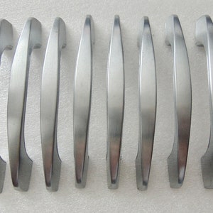 May include: A set of eight brushed nickel cabinet handles. Each handle has a curved, elongated design with a rectangular base and a smooth, matte finish. The handles are arranged in a row, showcasing their consistent shape and metallic sheen.