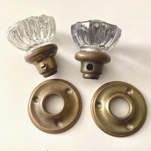 May include: Two vintage glass door knobs with brass bases and matching brass backplates. The clear glass knobs have a ribbed design. The brass components show signs of age and wear, adding to their antique charm.