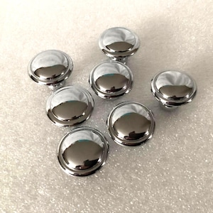 May include: Seven shiny, chrome-plated cabinet knobs with a rounded, domed design. The knobs are arranged on a textured, off-white surface, reflecting light and creating highlights. These hardware pieces are likely for furniture or cabinetry.