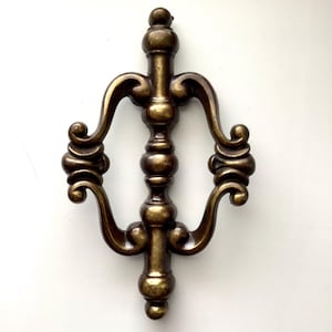 May include: Ornate, antique-style bronze metal door handle. The handle features a central vertical column with decorative curves and scrollwork on either side. The finish has a warm, aged appearance, suggesting vintage charm. The handle is designed for a door.