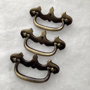 May include: Three antique brass drawer pulls with a decorative design. Each pull features a curved handle, ornate detailing, and a keyhole. The pulls are arranged in a vertical stack against a white background.