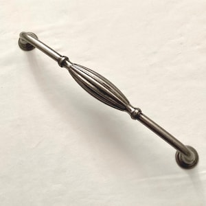May include: A dark gray cabinet pull with a decorative, ridged center section and rounded ends. The hardware is made of metal and has a vintage or antique appearance. The pull is designed for use on drawers or cabinet doors.