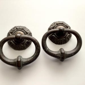 May include: Two antique bronze-colored door pulls. Each pull features a circular backplate with a decorative floral pattern and a rounded handle. The handles are oval-shaped and attached to the backplates. The hardware is likely for a cabinet or drawer.