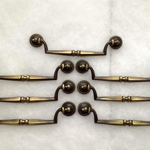 May include: A set of seven matching antique brass drawer pulls arranged in a grid. Each pull features a central horizontal bar with a decorative ribbed detail, connected to two curved arms that terminate in circular mounting plates with small knobs.