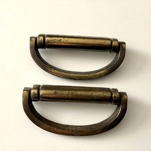 May include: Two antique brass drawer pulls with a curved handle design. The pulls have a cylindrical center and a slightly aged, tarnished appearance, suggesting vintage or reclaimed hardware. Ideal for furniture restoration projects.