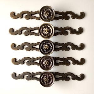May include: Five antique bronze cabinet pulls. Each pull features a circular floral design in the center, with ornate scrollwork on either side. The pulls are arranged in a row against a white background. Hardware for furniture.
