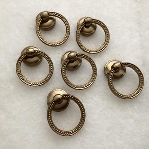 May include: A set of six circular brass-toned metal drawer pulls. Each pull features a round mounting base and a hinged ring handle with a beaded texture along the outer edge. The hardware has a matte finish and a consistent metallic color.