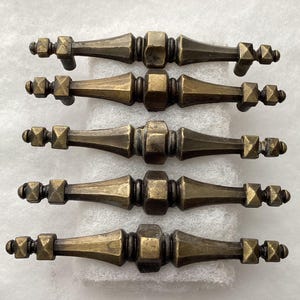 May include: Five antique bronze cabinet pulls. Each pull features a central hexagonal element flanked by decorative diamond-shaped accents and small, rounded end pieces. The pulls are arranged horizontally, showcasing their detailed design and aged finish.
