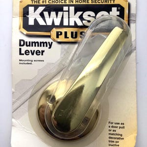 May include: A Kwikset Plus dummy lever in its original packaging. The lever is gold-colored and has a curved design. The packaging includes the text "Dummy Lever" and "Mounting screws included."