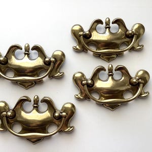 May include: Four antique brass drawer pulls with a curved handle and ornate design. The pulls have a warm, golden hue and are arranged on a white surface. Each pull measures approximately 4 inches wide.