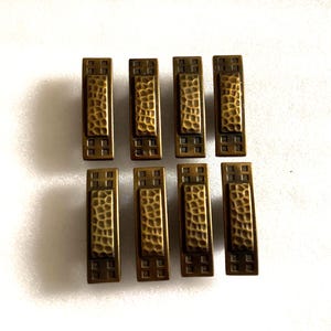 May include: A set of eight rectangular cabinet pulls in an antique brass finish. Each pull features a central panel with a hammered texture, flanked by geometric square patterns at the top and bottom ends, arranged in two rows of four pieces.