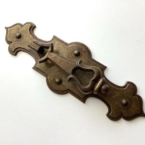 May include: Ornate antique bronze-toned cabinet pull with a decorative handle and a rectangular backplate. The pull features a keyhole and is embellished with rounded rivets and a scrolled design, suitable for vintage furniture.