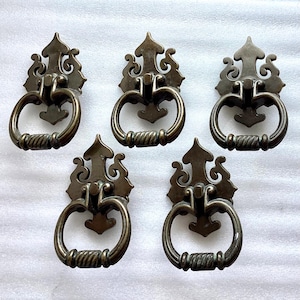 May include: Five antique brass cabinet pulls with ornate designs. Each pull features a decorative, scrolled top and a ring-shaped handle.