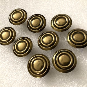 May include: A collection of ten antique brass cabinet knobs. Each knob is round with concentric circles, creating a layered effect. The knobs have a warm, aged brass finish, suggesting vintage or retro design.