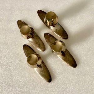 May include: Four vintage gold-toned collar stays with a rounded top and an elongated, pointed base. The collar stays are arranged on a white surface, showcasing their aged, tarnished appearance. These accessories are designed to keep a shirt collar in place.