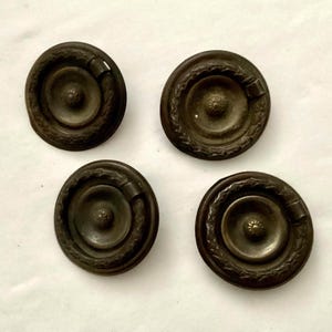 May include: Four antique bronze-toned drawer pulls. Each pull is circular with a central knob and a decorative outer ring. The pulls have a dark, aged appearance, suggesting vintage or vintage-inspired hardware.