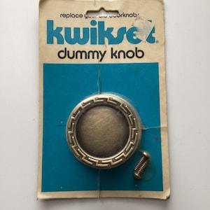 May include: A vintage Kwikset dummy knob replacement kit. The packaging is a worn, off-white card with blue and teal accents. The knob is a circular, bronze-colored metal with a decorative border. Includes a single screw.