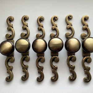 May include: Set of six antique brass cabinet pulls. Each pull features a round knob and an ornate, curved backplate. The brass has a warm, aged patina, suggesting vintage or retro design. Ideal for furniture restoration or adding a classic touch.