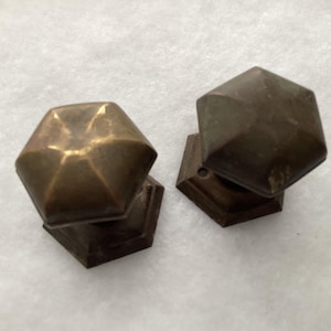 May include: Two hexagonal brass door knobs with a weathered, dark metallic finish. Each knob features a six-sided geometric top and a matching hexagonal base plate, showing signs of oxidation and patina across the surface of the metal.
