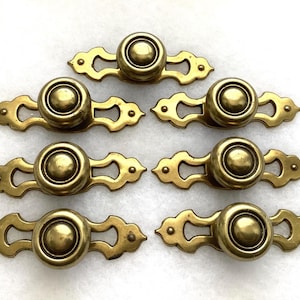 May include: A set of six brass drawer pulls, each featuring a central circular knob mounted on an elongated, decorative backplate with scalloped edges and pointed ends. The metal has a polished, golden-toned finish throughout.