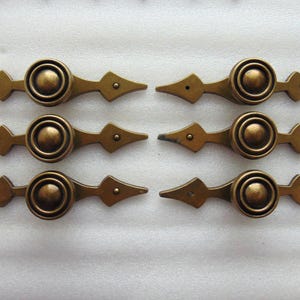 May include: Six antique brass drawer pulls. Each pull features a central circular design with a raised center, flanked by diamond-shaped accents and pointed ends. The pulls have a warm, aged patina.