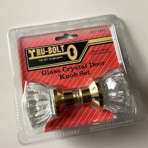 May include: A glass crystal door knob set in a clear plastic package. The knobs are clear with a ribbed design, and the base is gold-colored. The packaging has red and white accents with the text "TRU-BOLT" and "Glass Crystal Door Knob Set."
