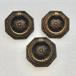 May include: Three antique bronze-colored drawer pulls with a decorative octagonal shape. Each pull features a central floral design and a ring handle. The pulls are likely for furniture restoration or decorative use.