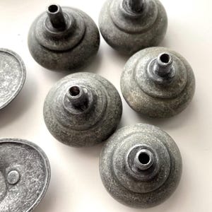 May include: A selection of grey cabinet knobs and pulls with a weathered finish. The knobs are round with a tiered design and a central hole. The pulls are oval-shaped with a single hole. The hardware has a rustic, antique appearance.