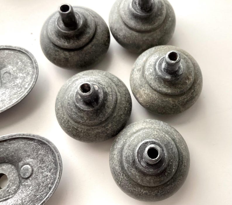 May include: A selection of grey cabinet knobs and pulls with a weathered finish. The knobs are round with a tiered design and a central hole. The pulls are oval-shaped with a single hole. The hardware has a rustic, antique appearance.