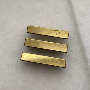 May include: Three rectangular, gold-colored metal bars are arranged horizontally. The bars have a slightly worn appearance, suggesting they may be vintage or have been used. The background is a plain, off-white surface.