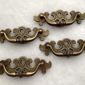 May include: Four brass-toned metal drawer pulls featuring a decorative backplate with an intricate cutout design and a curved, swinging handle. Each piece has a weathered finish and is arranged in a scattered pattern on a light surface.