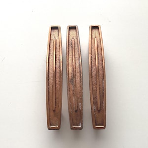 May include: Three vintage copper-toned cabinet pulls. Each pull has a rectangular shape with a central groove and a slightly curved design. The pulls have a distressed finish, showing signs of age and use. They are arranged on a white surface.